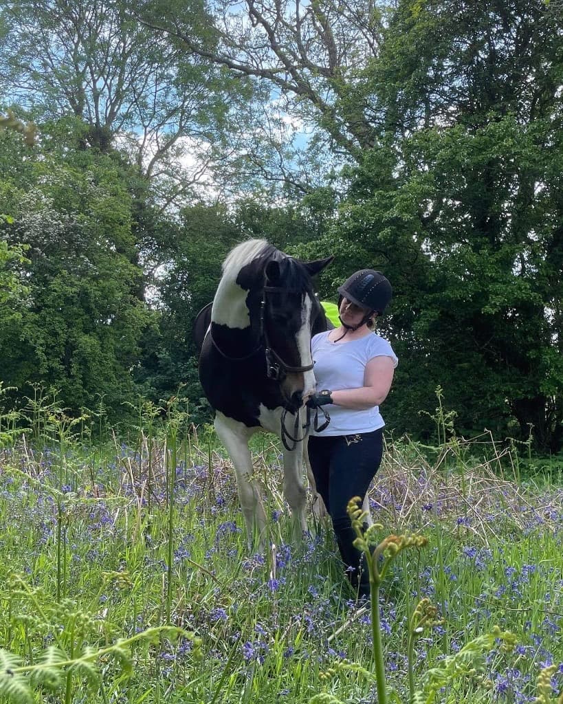 Lyndsey standing with her black-and-white horse in a woodland meadow